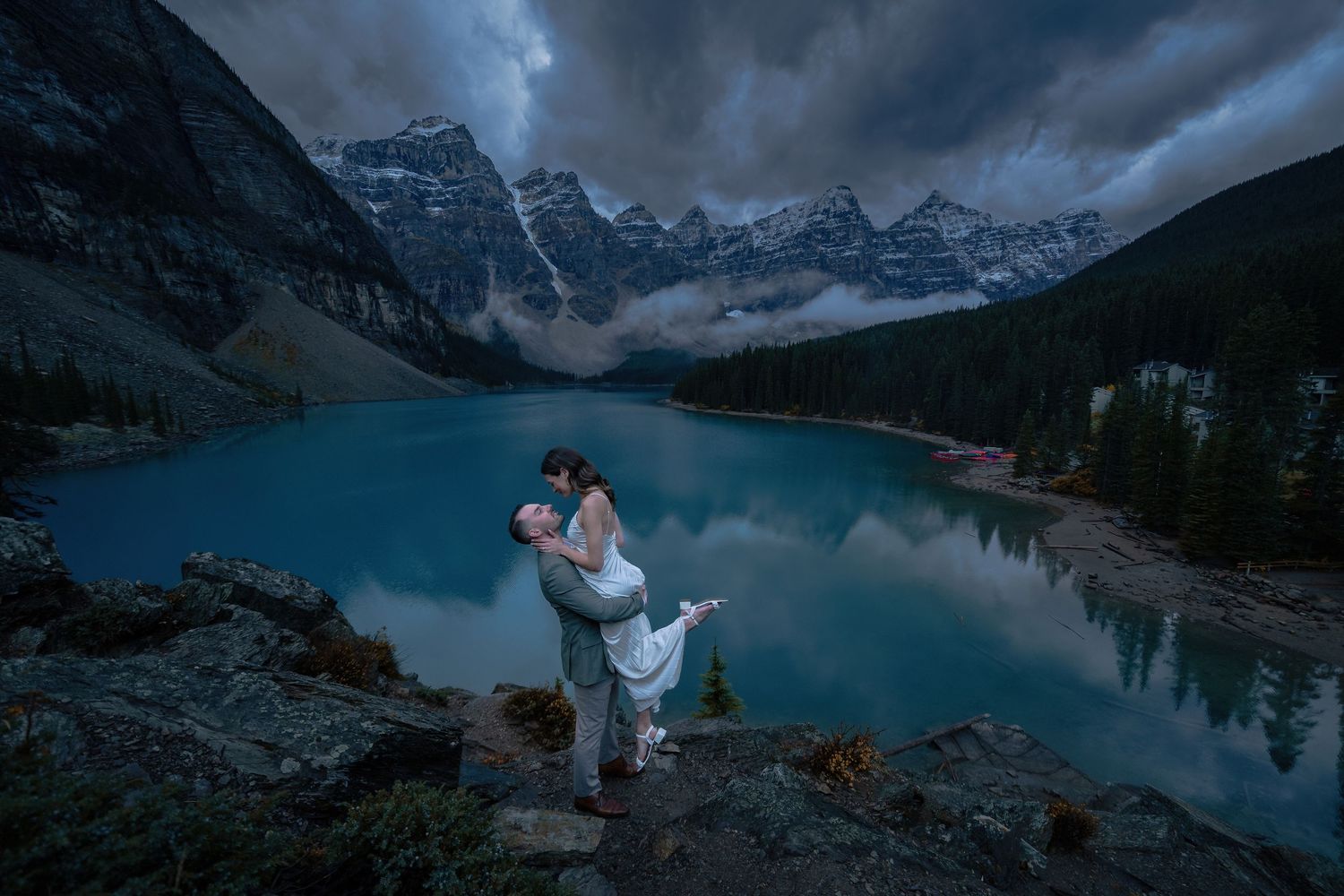 Couple shares romantic embrace at Moraine Lake, Banff, with turquoise waters and dramatic mountain peaks under stormy skies.