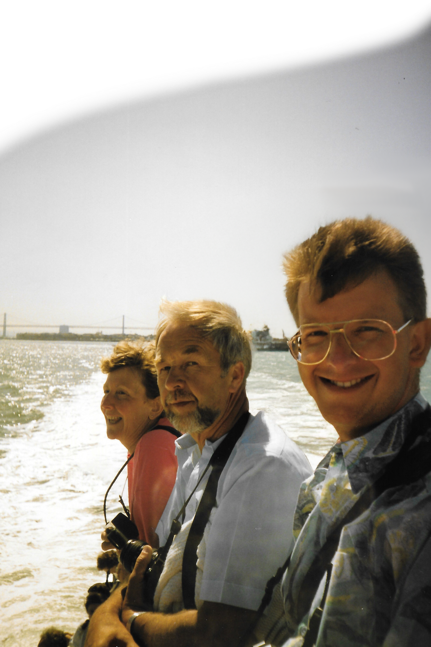 Three smiling people on boat with choppy water and bridge visible in sunny background.