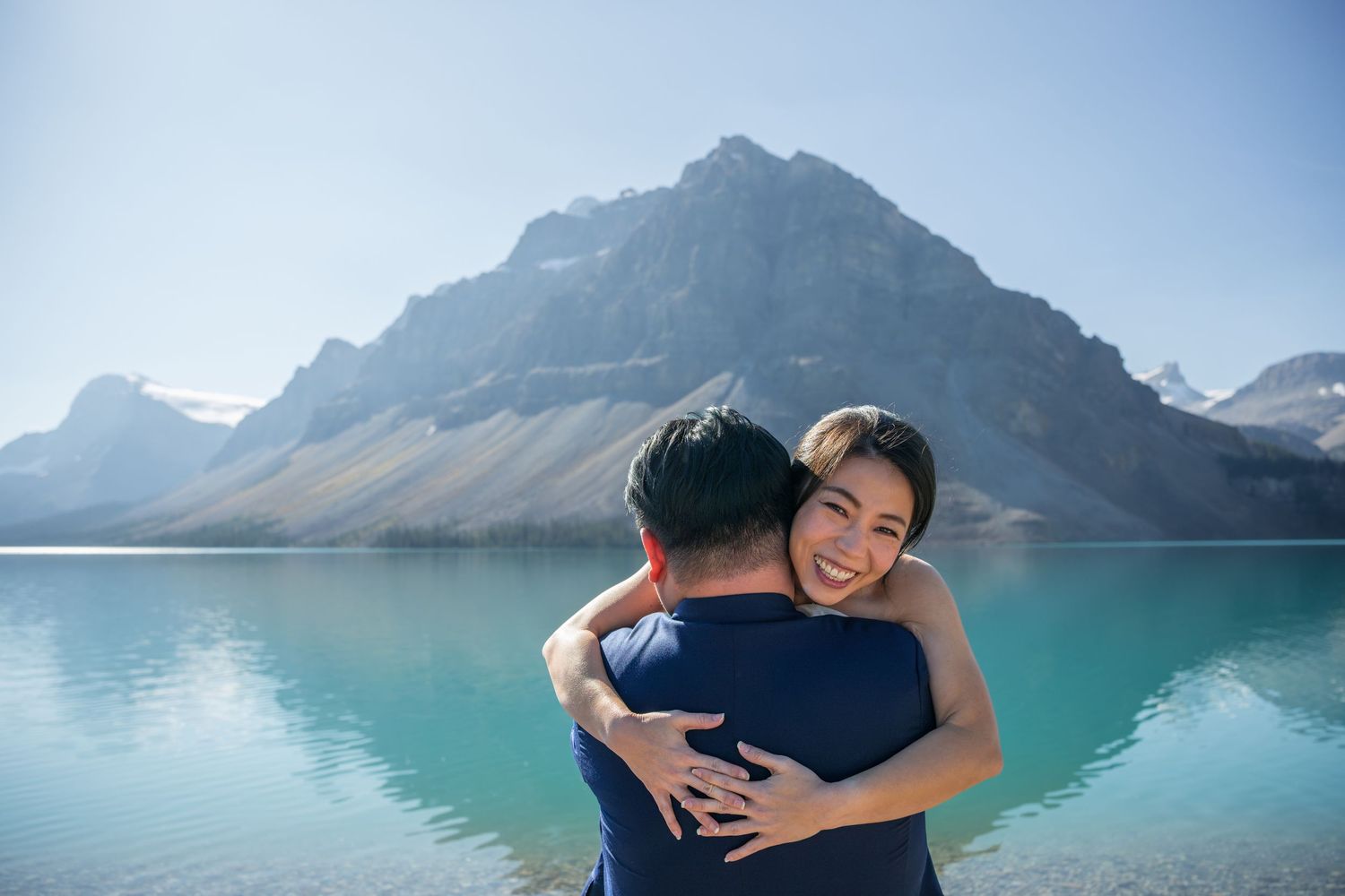 Couple embracing and smiling by a turquoise mountain lake under a clear blue sky.
