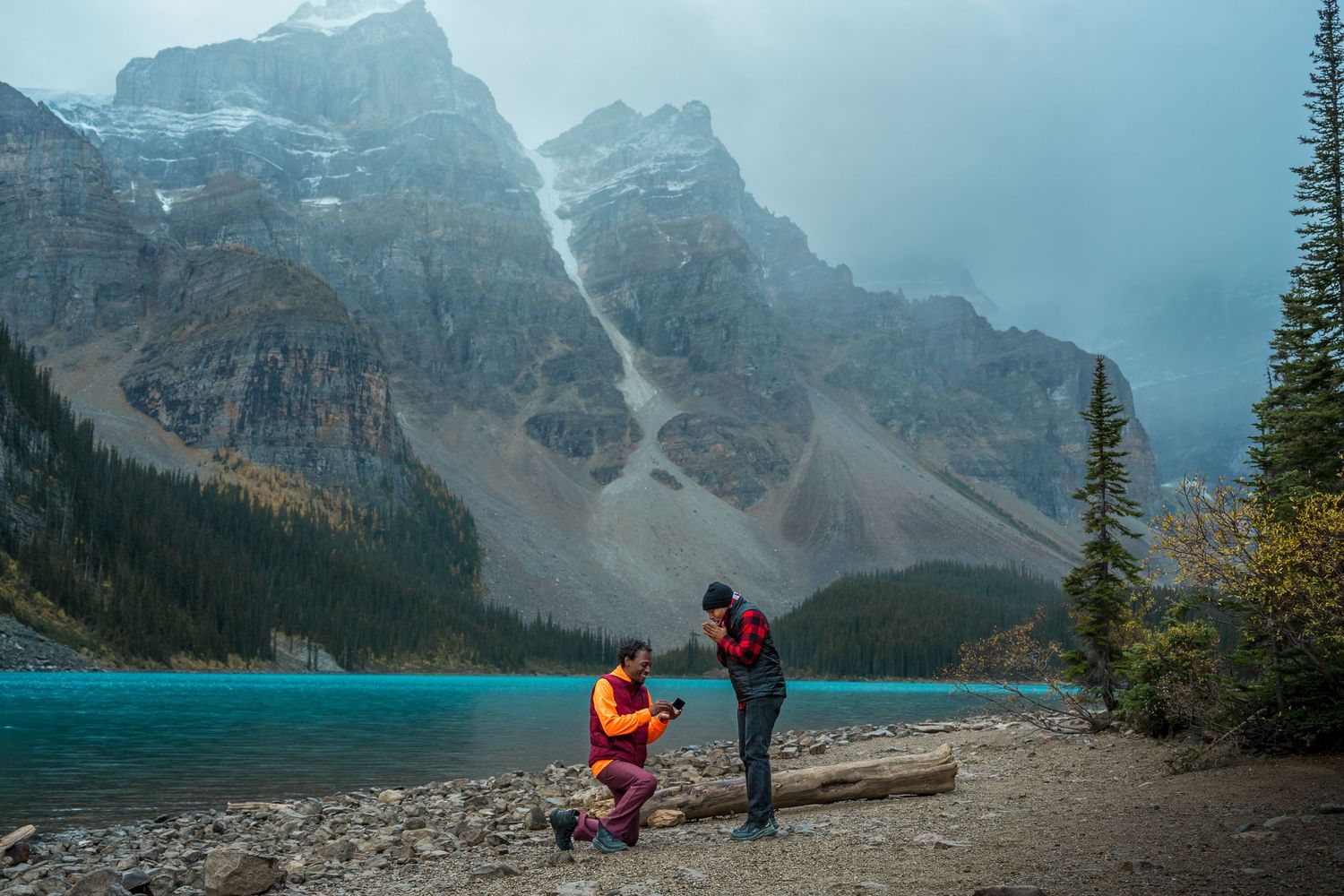 Couple sharing a proposal moment by turquoise Moraine Lake with dramatic rocky mountains in Banff, Canada.