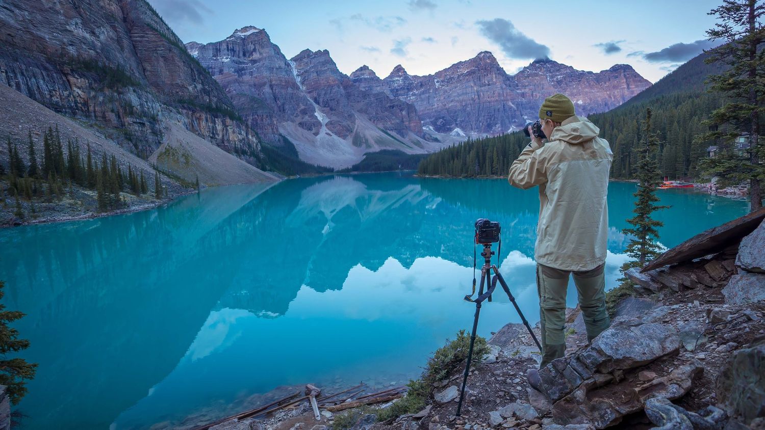 Photographer with tripod capturing stunning turquoise Moraine Lake at dusk with Rocky Mountain peaks behind.