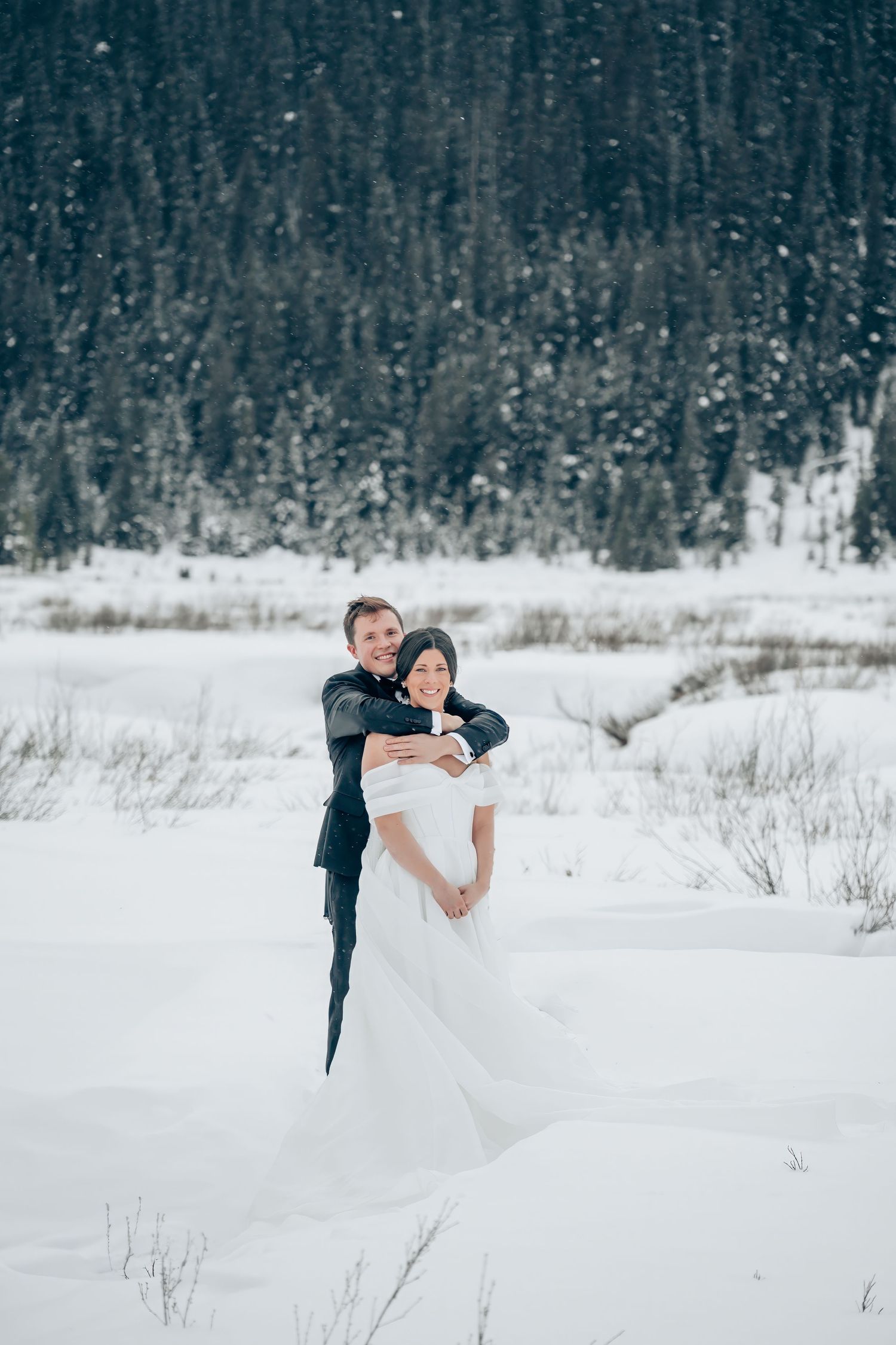 Bride and groom embracing in snowy winter landscape with snow-covered evergreen forest backdrop.
