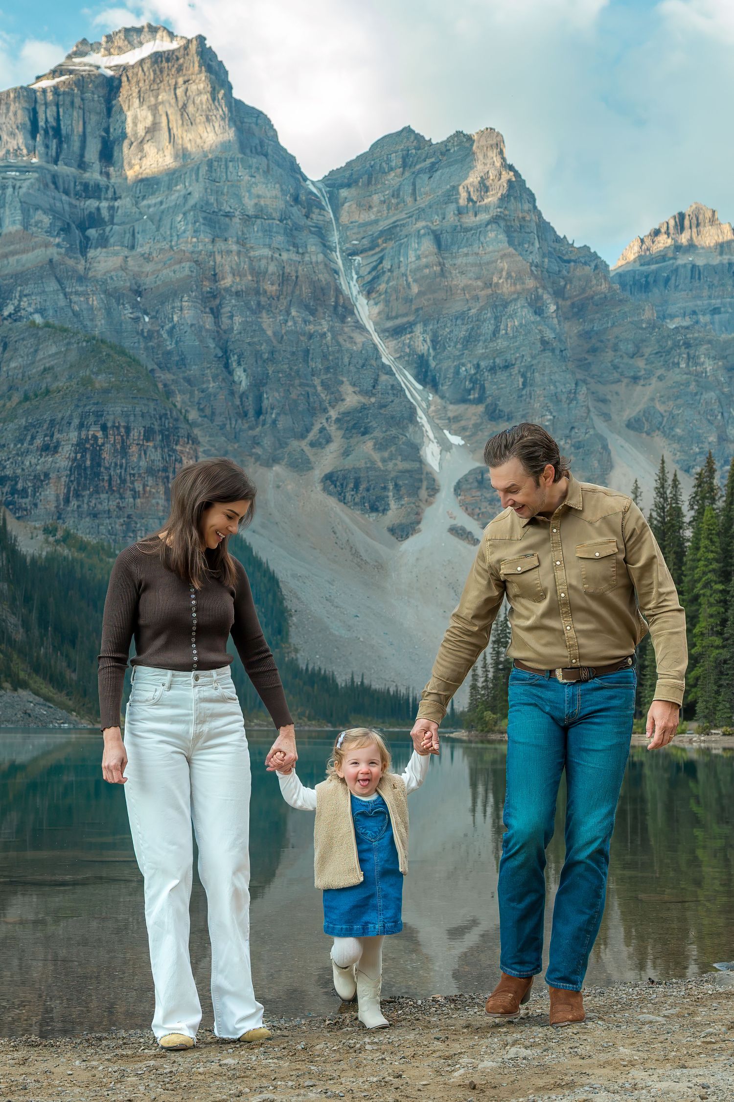 Family of three holding hands and smiling at Moraine Lake with towering Rocky Mountain peaks behind.