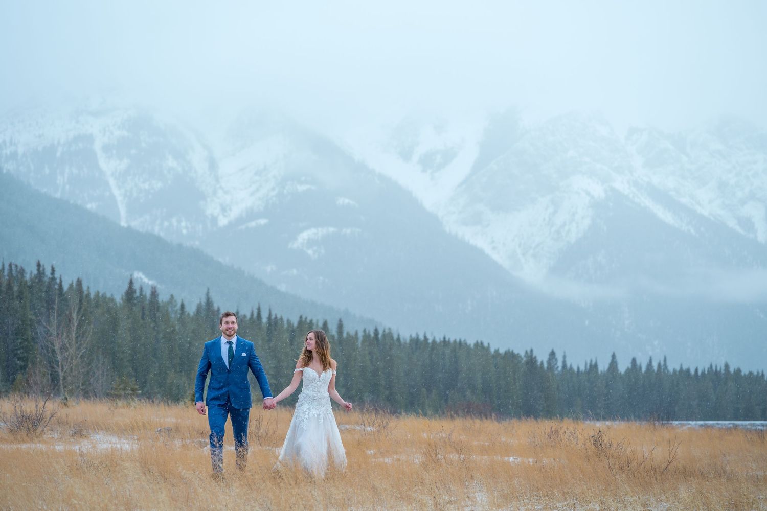 Bride and groom holding hands in snowy mountain landscape with golden grass and evergreen trees in background.