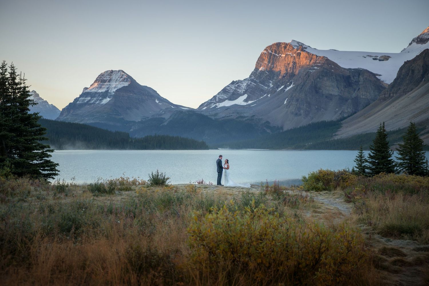 Couple standing by a serene mountain lake at sunset with snow-capped peaks in Banff National Park, Canada.