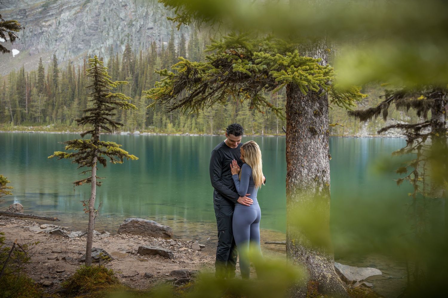 Couple embracing near turquoise alpine lake surrounded by pine trees and mountains in a romantic outdoor engagement photo.