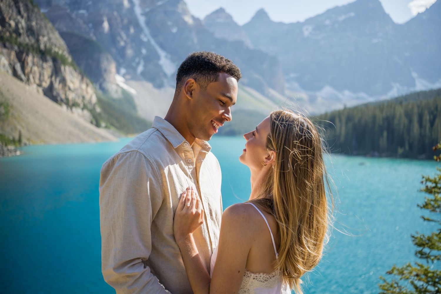 Couple embracing and smiling at each other with turquoise Moraine Lake and Rocky Mountains in background.