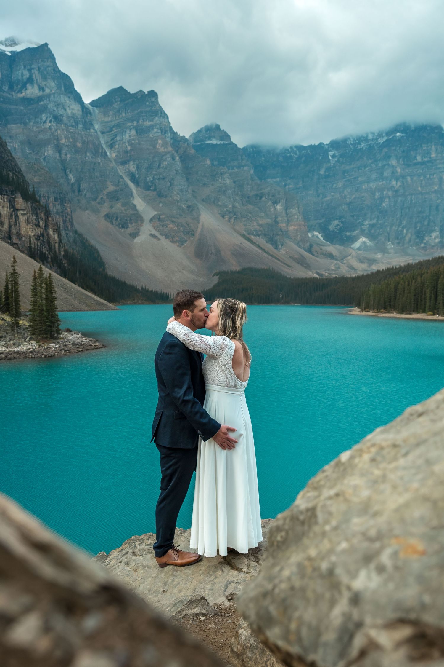 Couple kissing in wedding attire at Moraine Lake, Banff, with turquoise water and Rocky Mountains backdrop.
