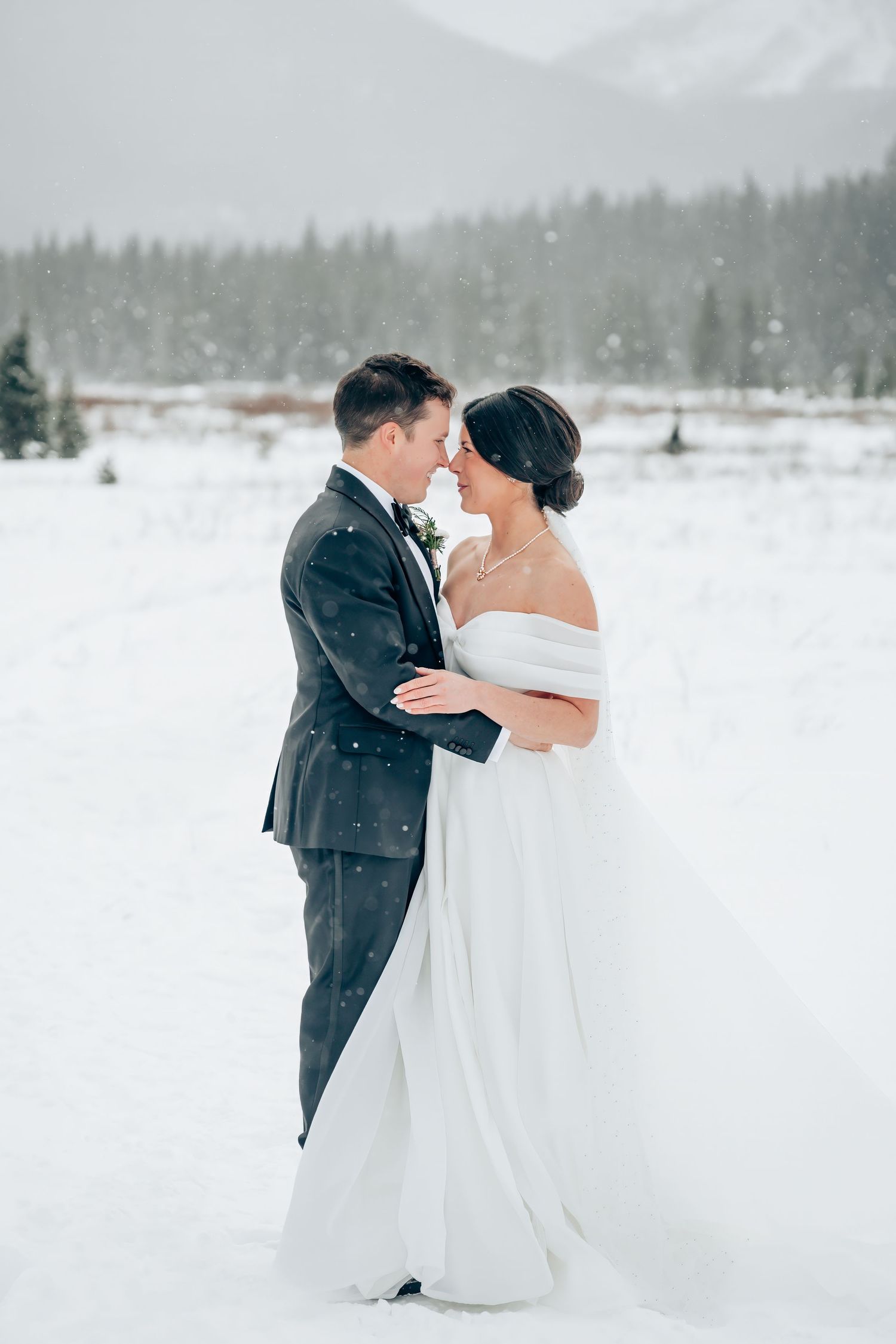 Bride and groom embrace in snowy winter landscape, she wears off-shoulder white gown, he wears dark suit, mountains behind.
