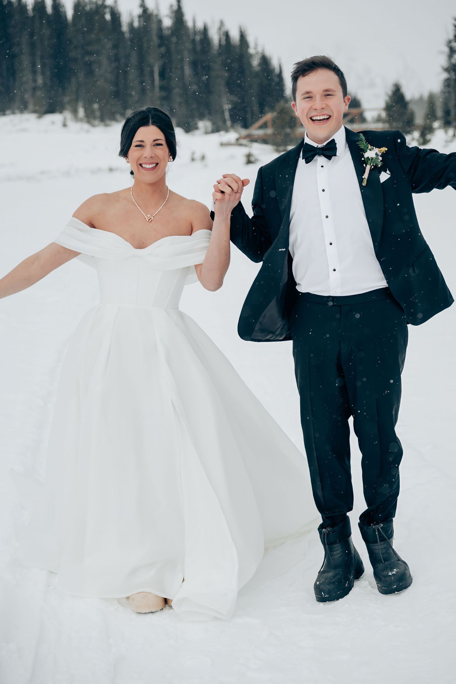Joyful bride in white off-shoulder gown and groom in black tuxedo holding hands, laughing in snowy winter wonderland.