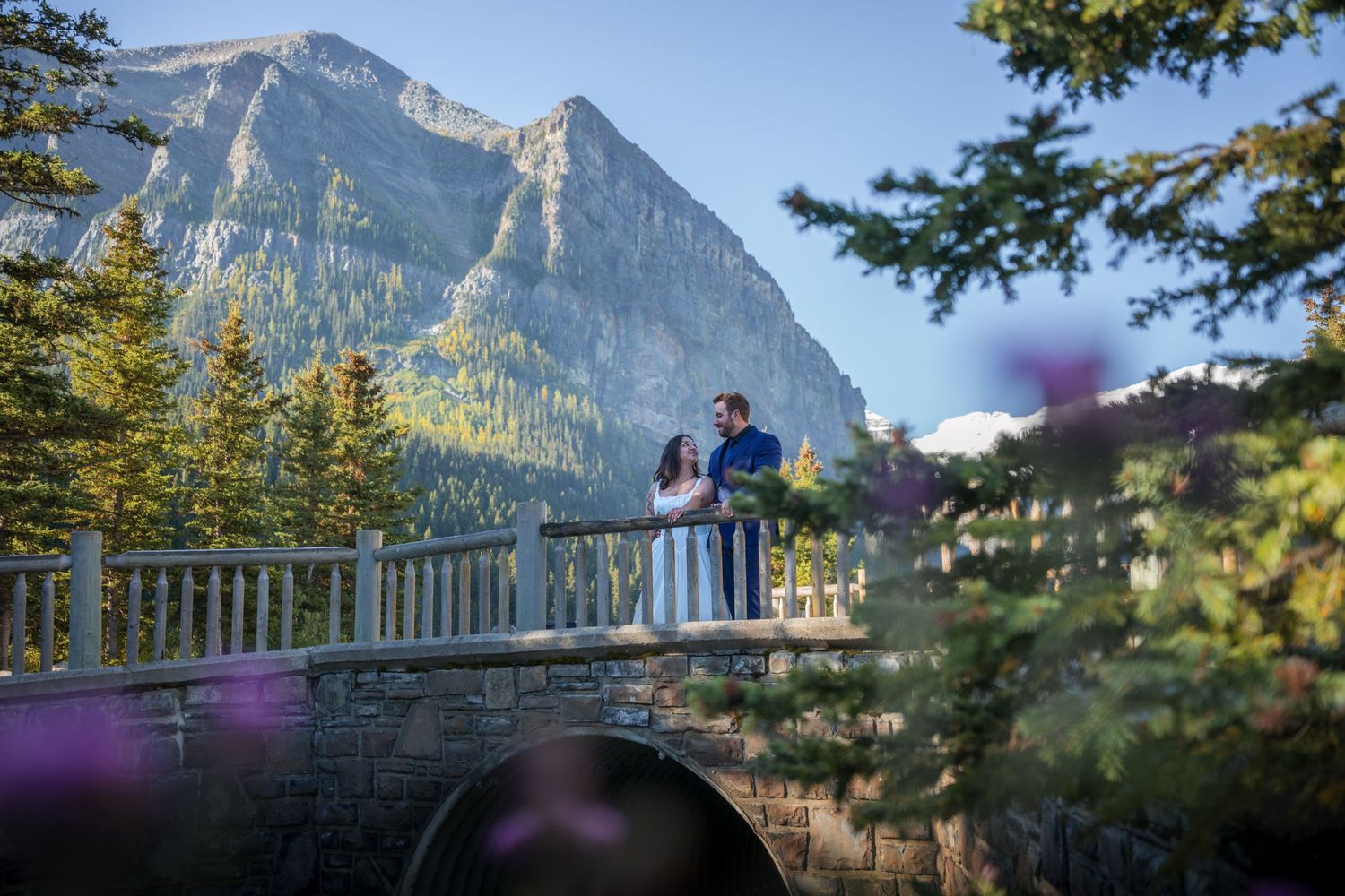 Couple standing on a stone bridge surrounded by pine trees with a majestic mountain backdrop at Lake Louise.