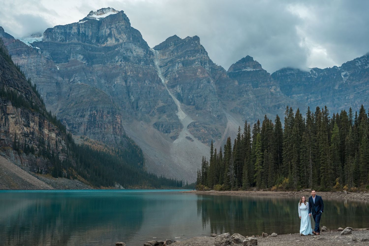 Wedding couple walks along shore of turquoise Moraine Lake with misty Rocky Mountains in Banff, Canada.