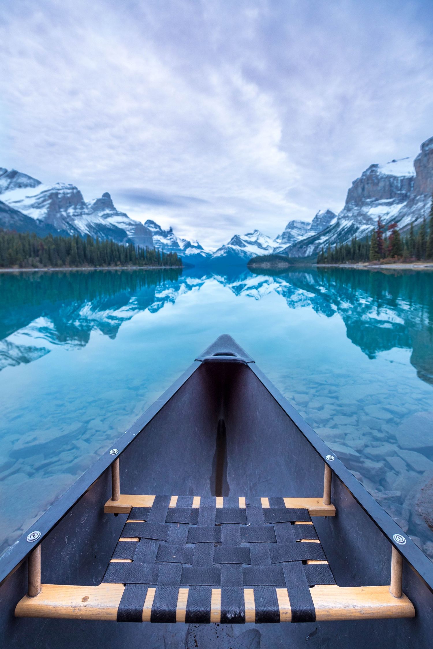 Wooden canoe bow pointing toward stunning turquoise glacial lake reflecting snow-capped mountains under cloudy sky.