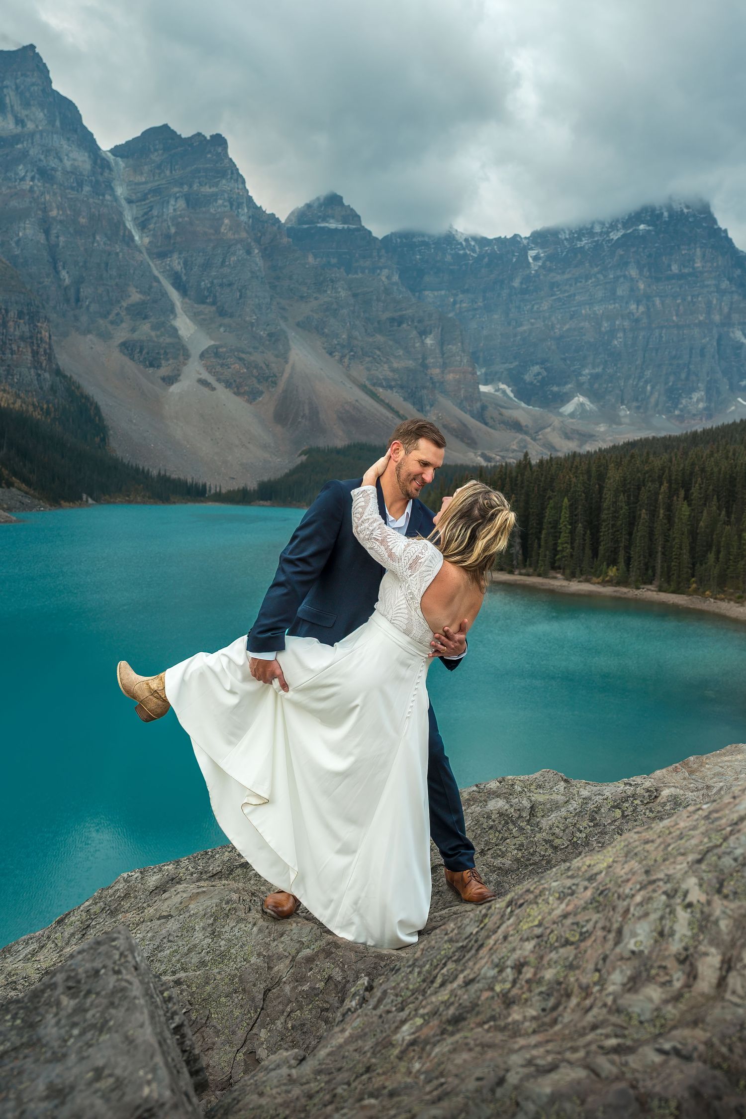 Bride and groom dancing on rocky overlook at Moraine Lake with dramatic stormy mountain backdrop.