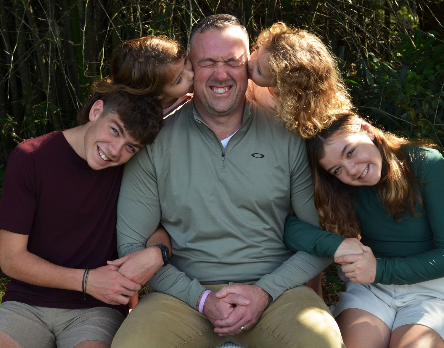 A cheerful family of four poses outdoors, with three children hugging and kissing a smiling man seated in the center.