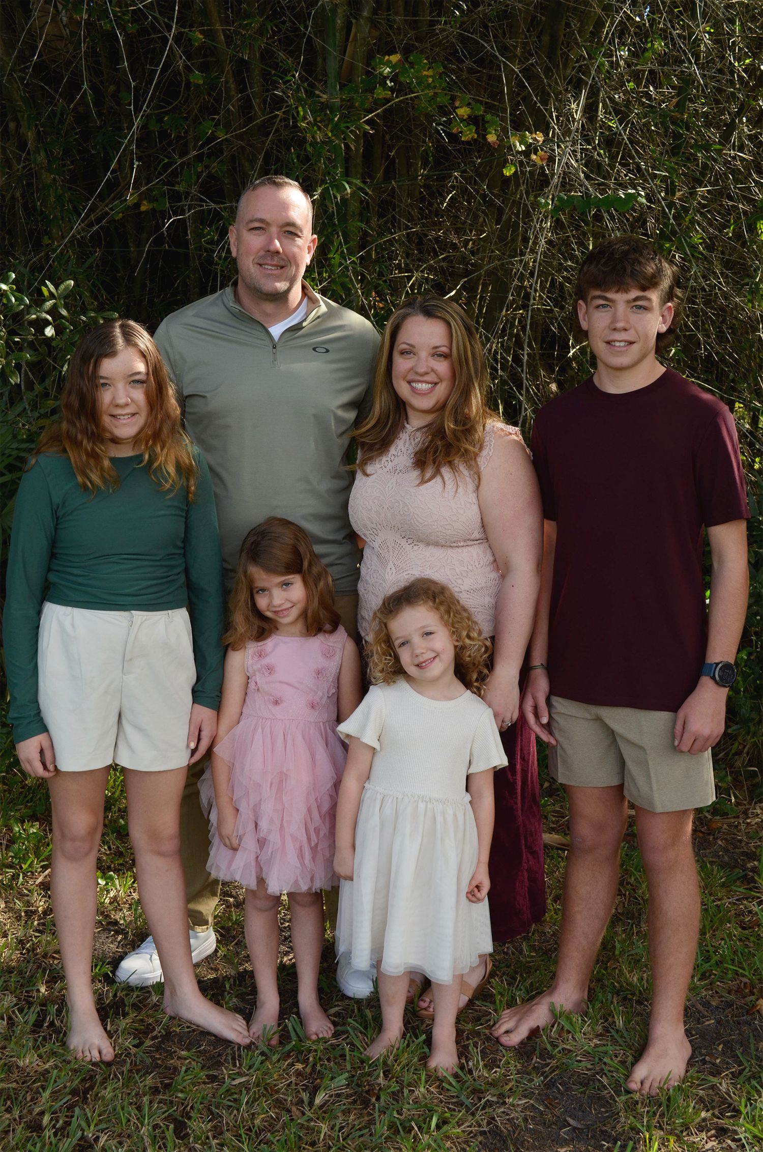 Family of six posing outdoors, parents with four children dressed in coordinating outfits, smiling together near lush greenery.