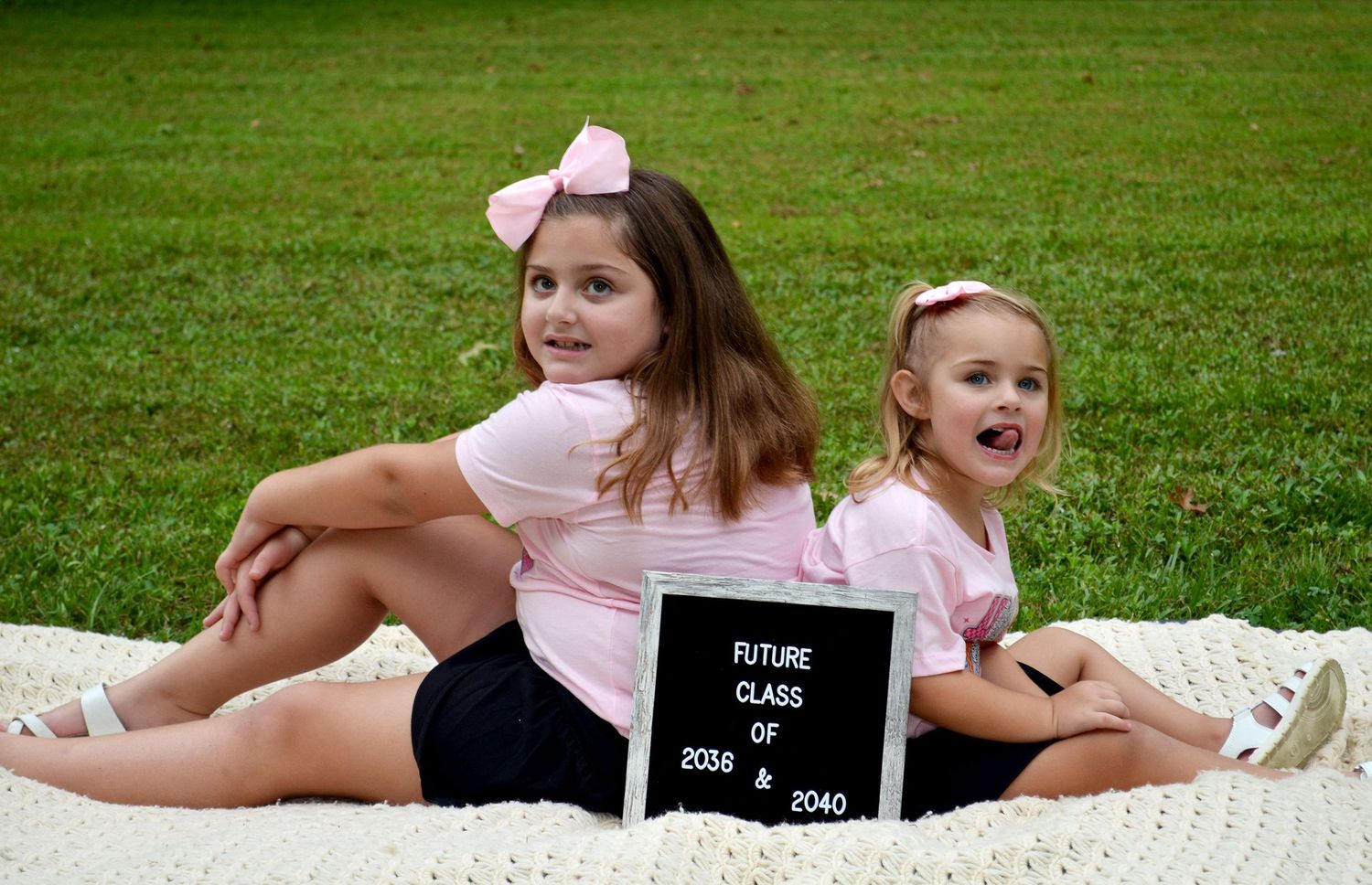 Two young girls in pink shirts sit back-to-back on a blanket holding a Future Class of 2036 and 2040 sign.
