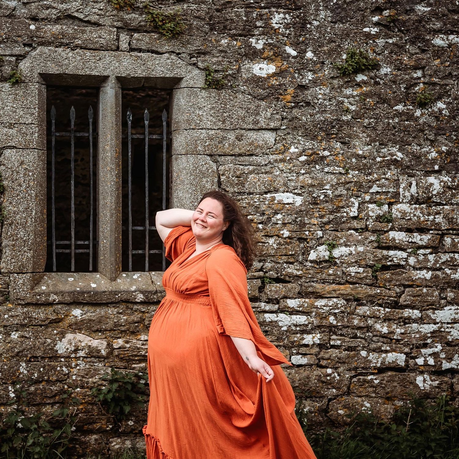 Woman laughing beside stone wall in flowing dress.