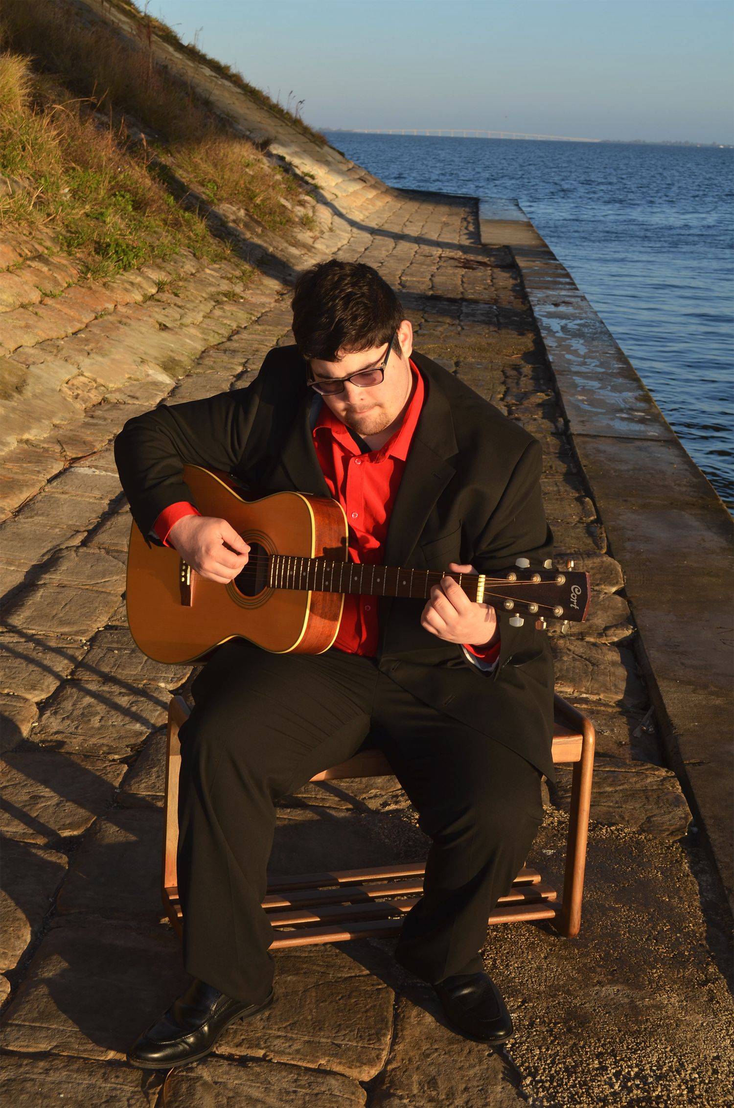 Teenage boy playing acoustic guitar seated by waterfront.