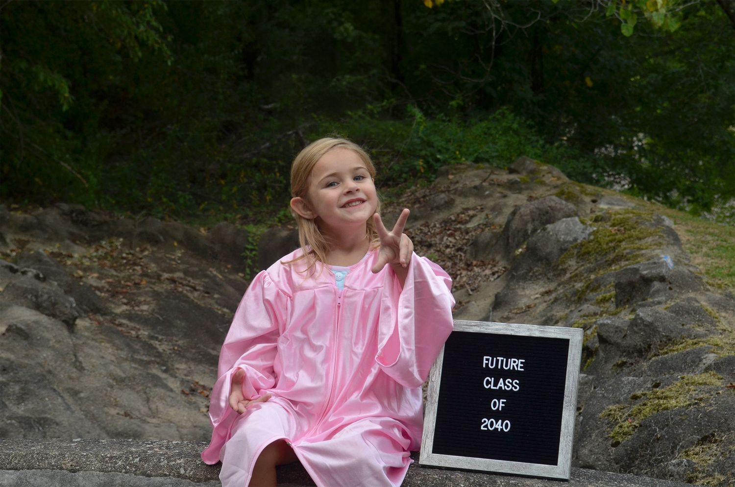 Young girl in graduation gown posing beside a future class sign on rocks.