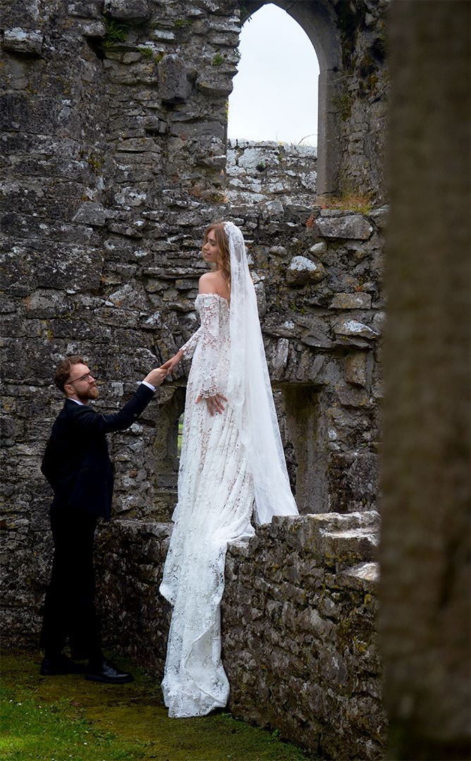 Bride standing in castle ruins while groom looks up at her.