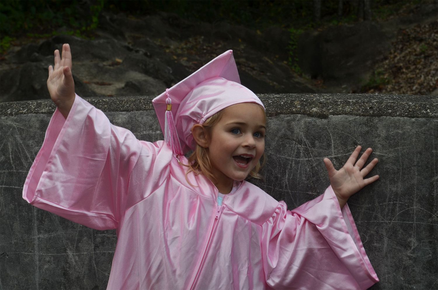 Excited young girl in pink graduation cap and gown celebrating with arms raised outdoors.