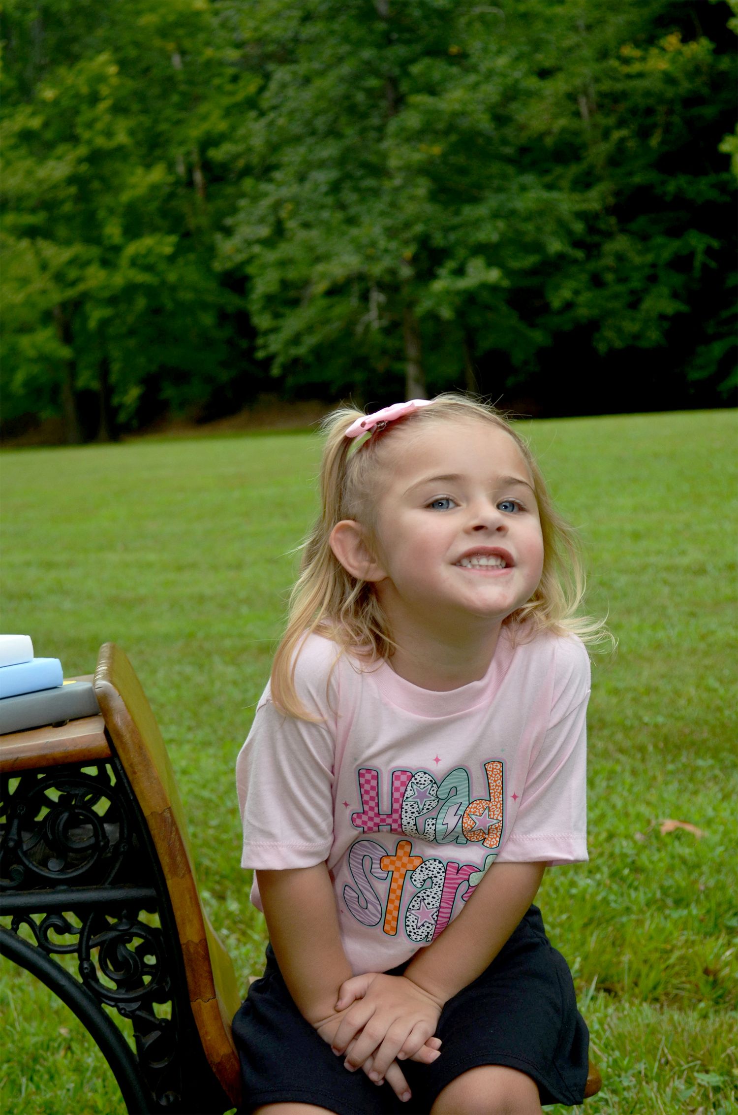 Young blonde girl in pink graphic tee smiling outdoors on green grass near a wooden chair.