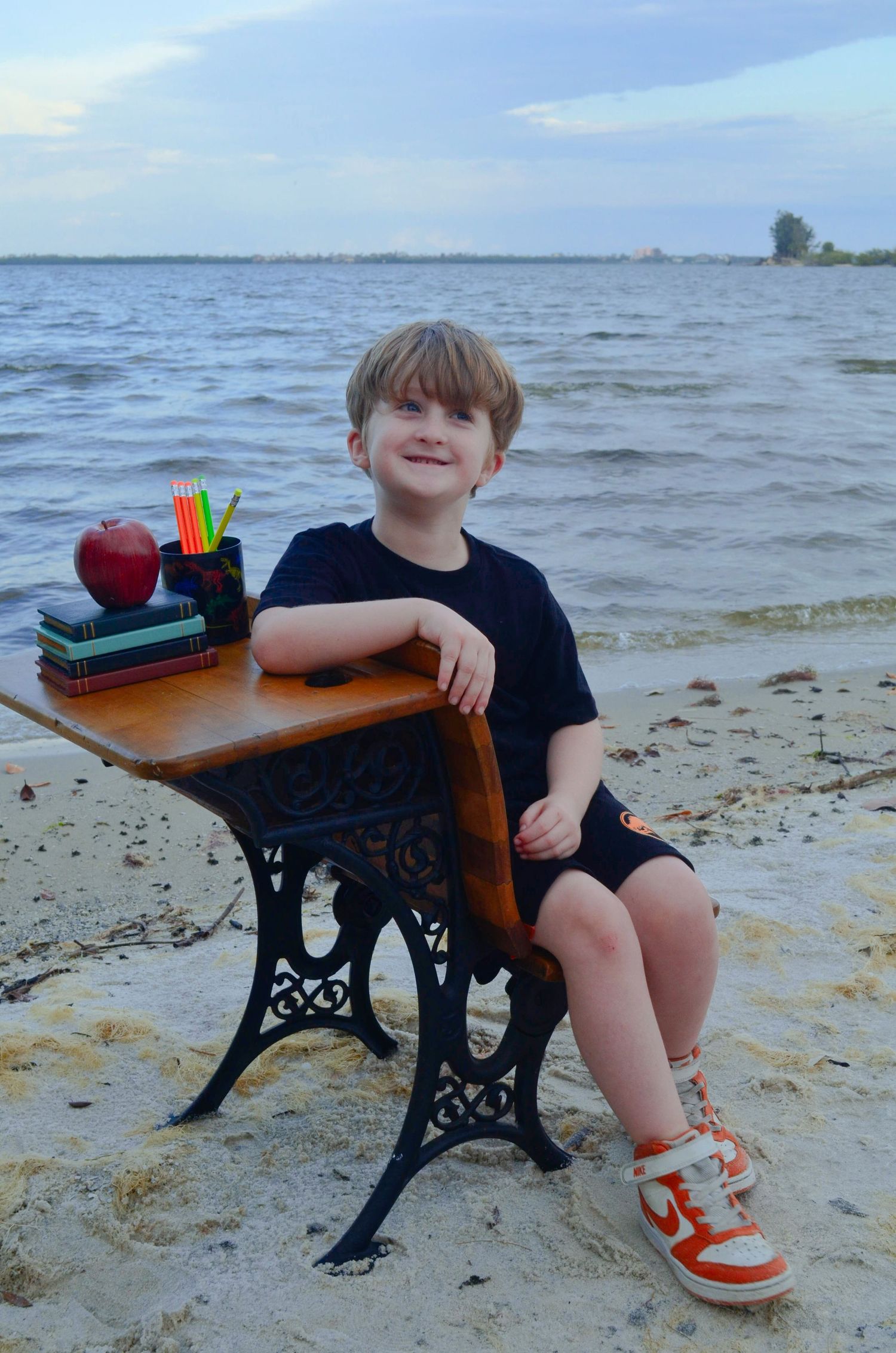 A smiling young boy sits at a vintage school desk on a sandy beach, with books, pencils, and an apple on the desk.