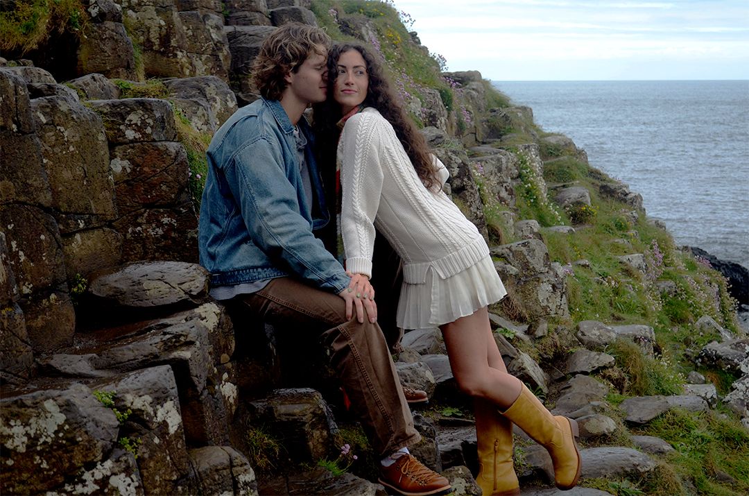 Couple posing romantically on ancient stone cliffs overlooking the sea, woman in white dress and yellow boots.