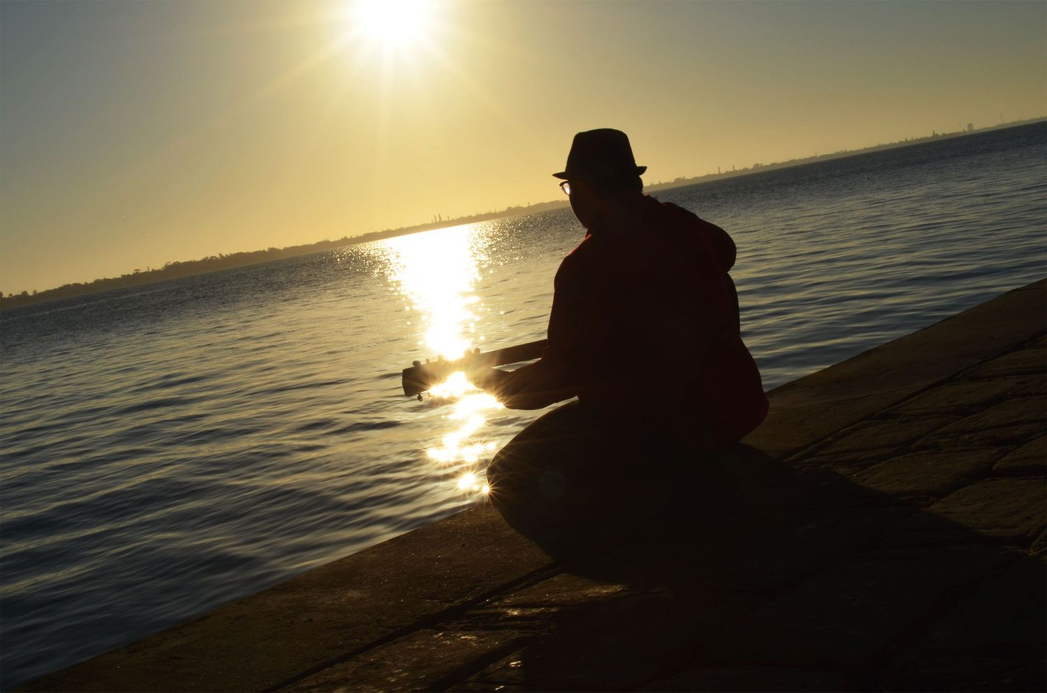 Silhouette of teenage boy sitting by water at sunrise.