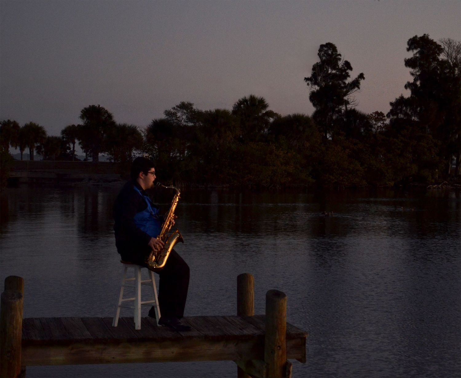 Teenage boy playing saxophone on wooden dock at dusk.