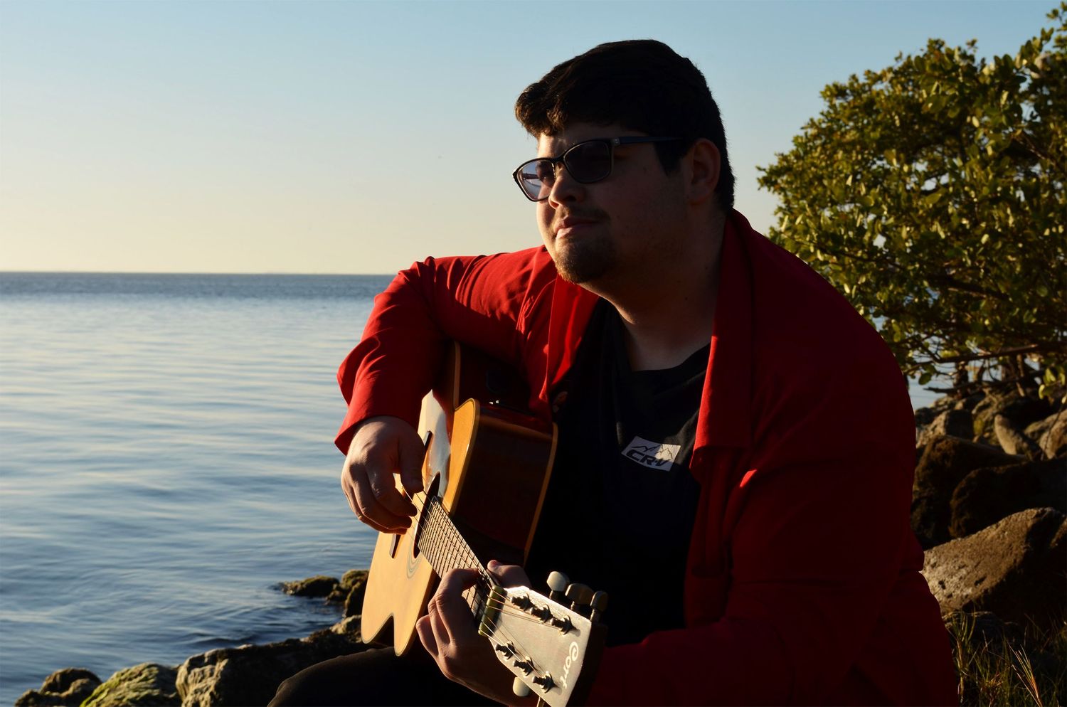 Teenage boy playing guitar on rocks by lake at sunrise.