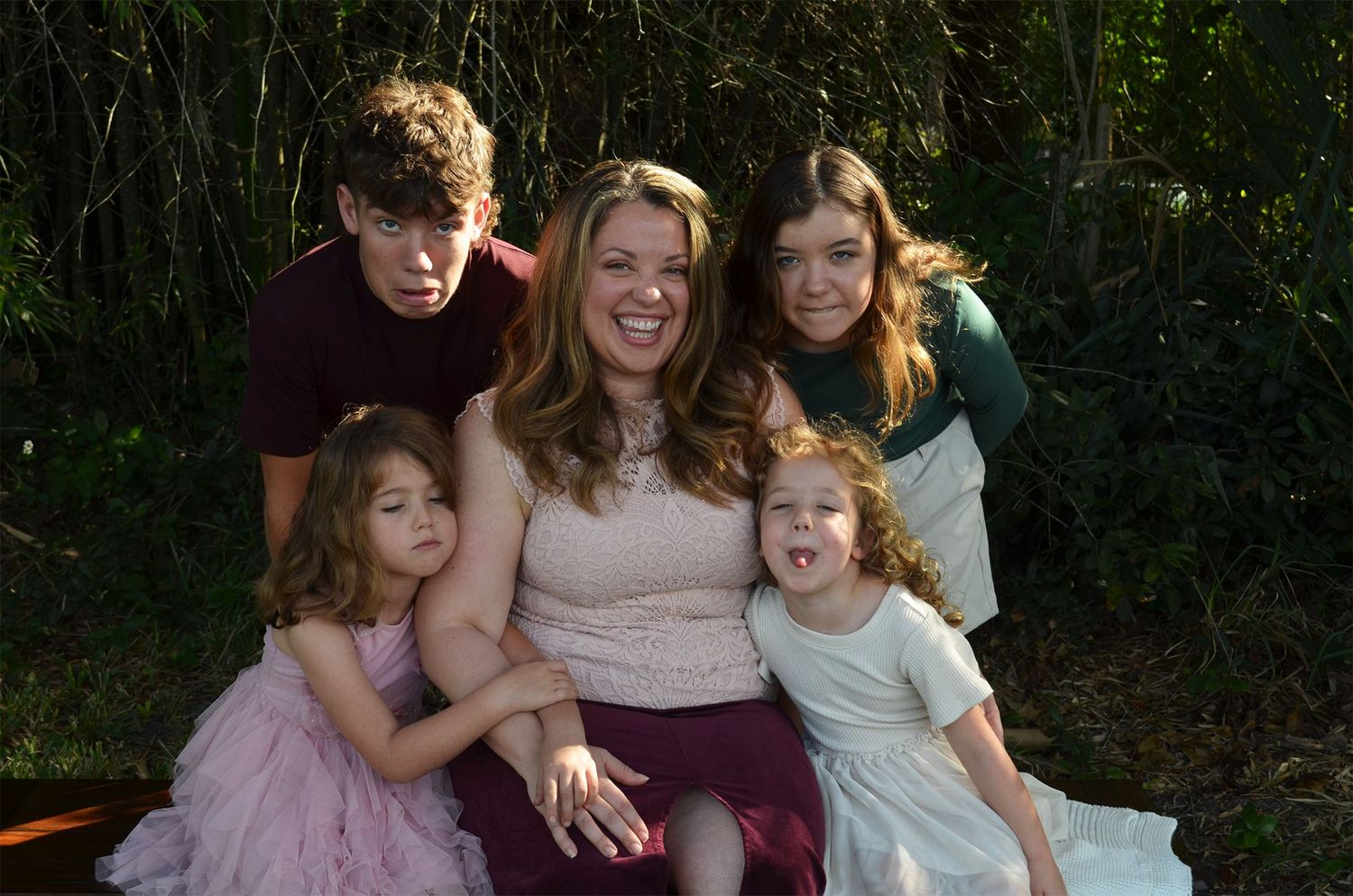 Family of five smiling together outdoors, with a mother and four children posing for a portrait in a garden setting.