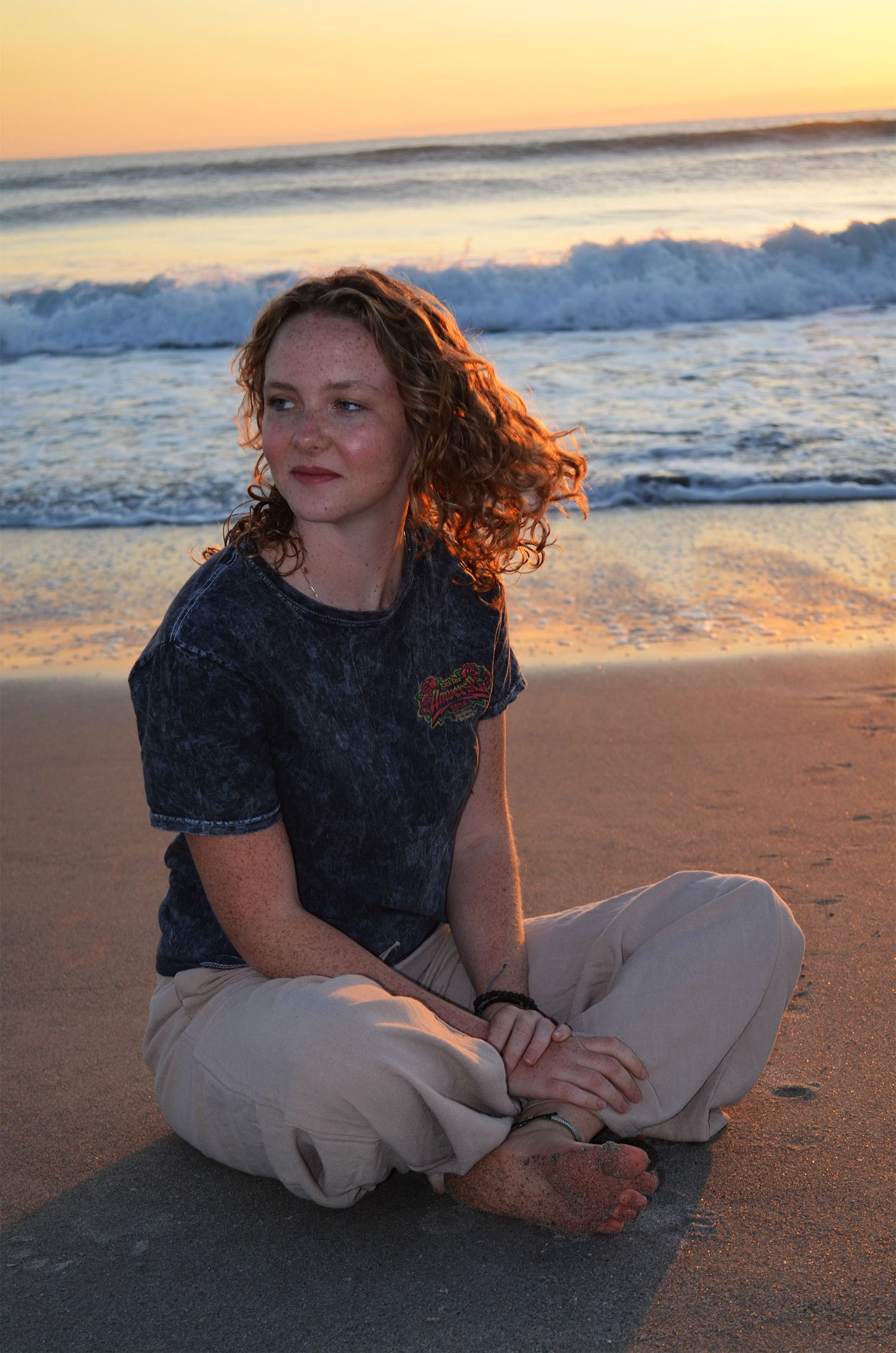 Teenage girl sitting cross-legged on beach at sunrise.