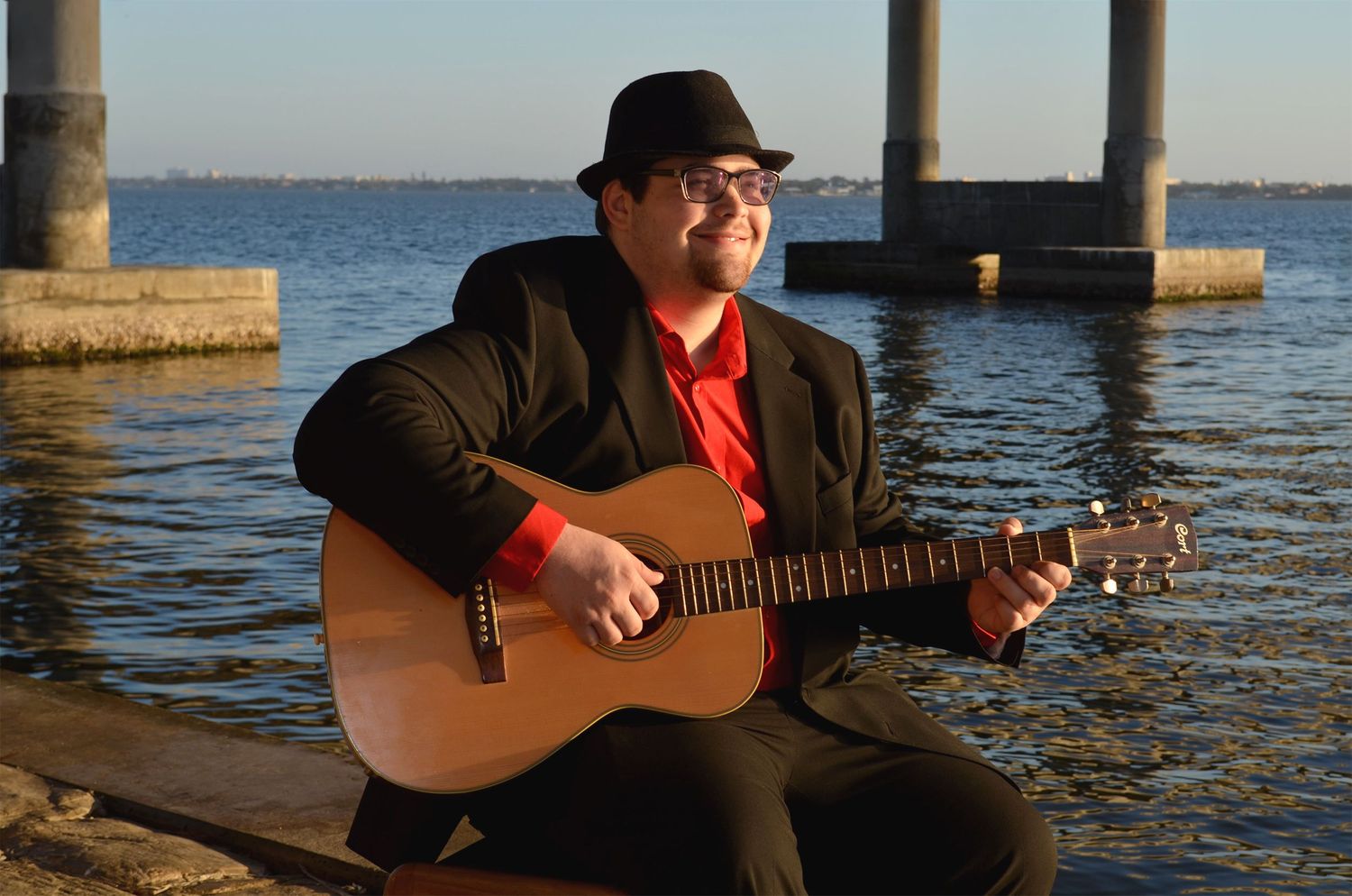 Teenage boy playing guitar near waterfront at sunrise.