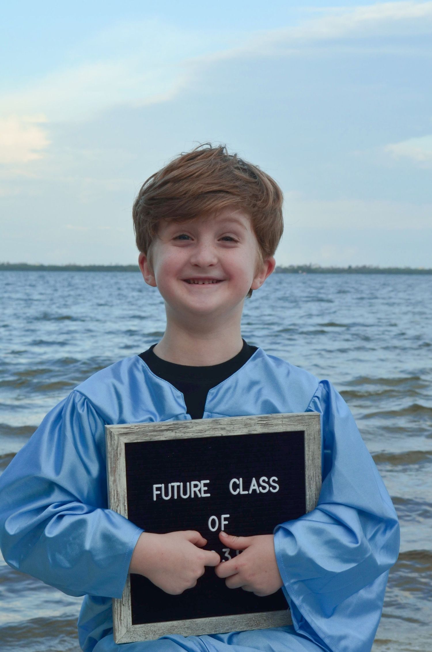 Boy in blue graduation gown holding future class sign near water.