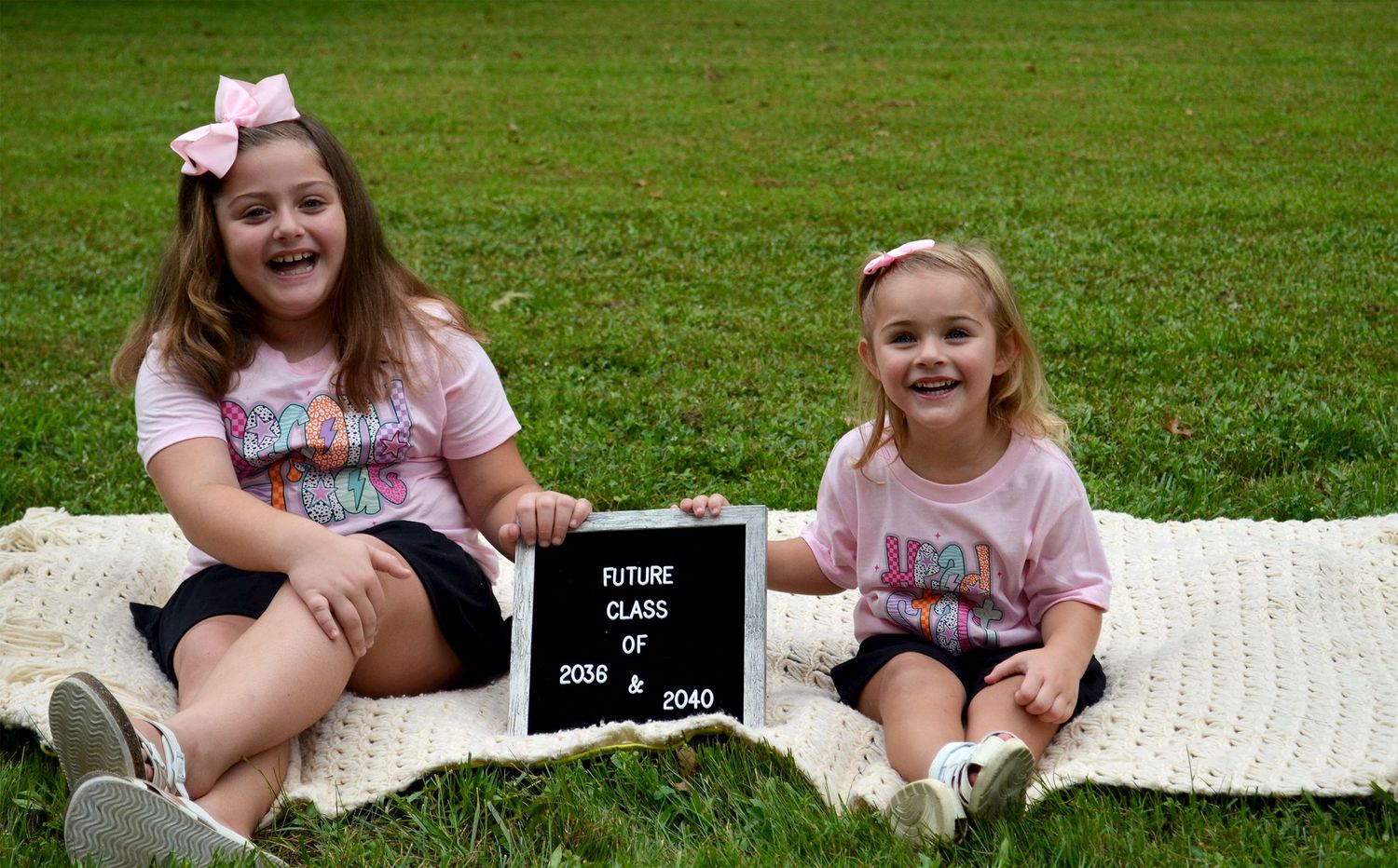Two sisters smiling while holding a future class letterboard on grass.