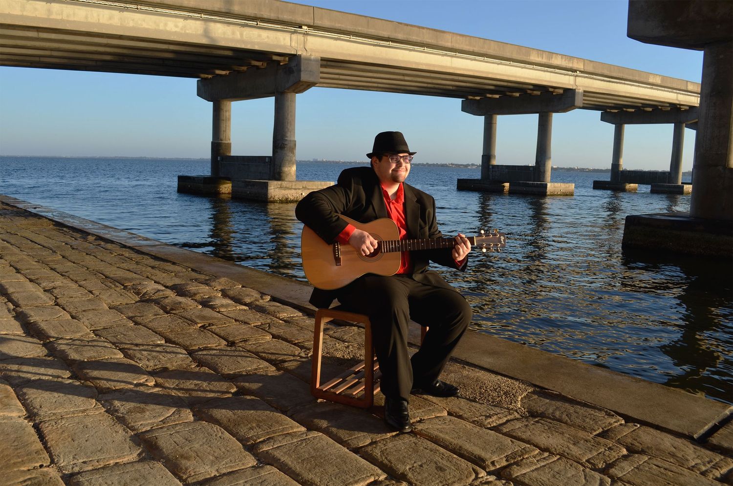 Teenage boy playing acoustic guitar on stone pier at sunrise.
