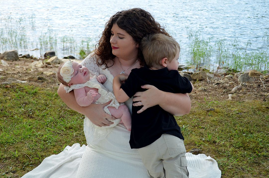 Mother holding newborn while son hugs her beside a lake.