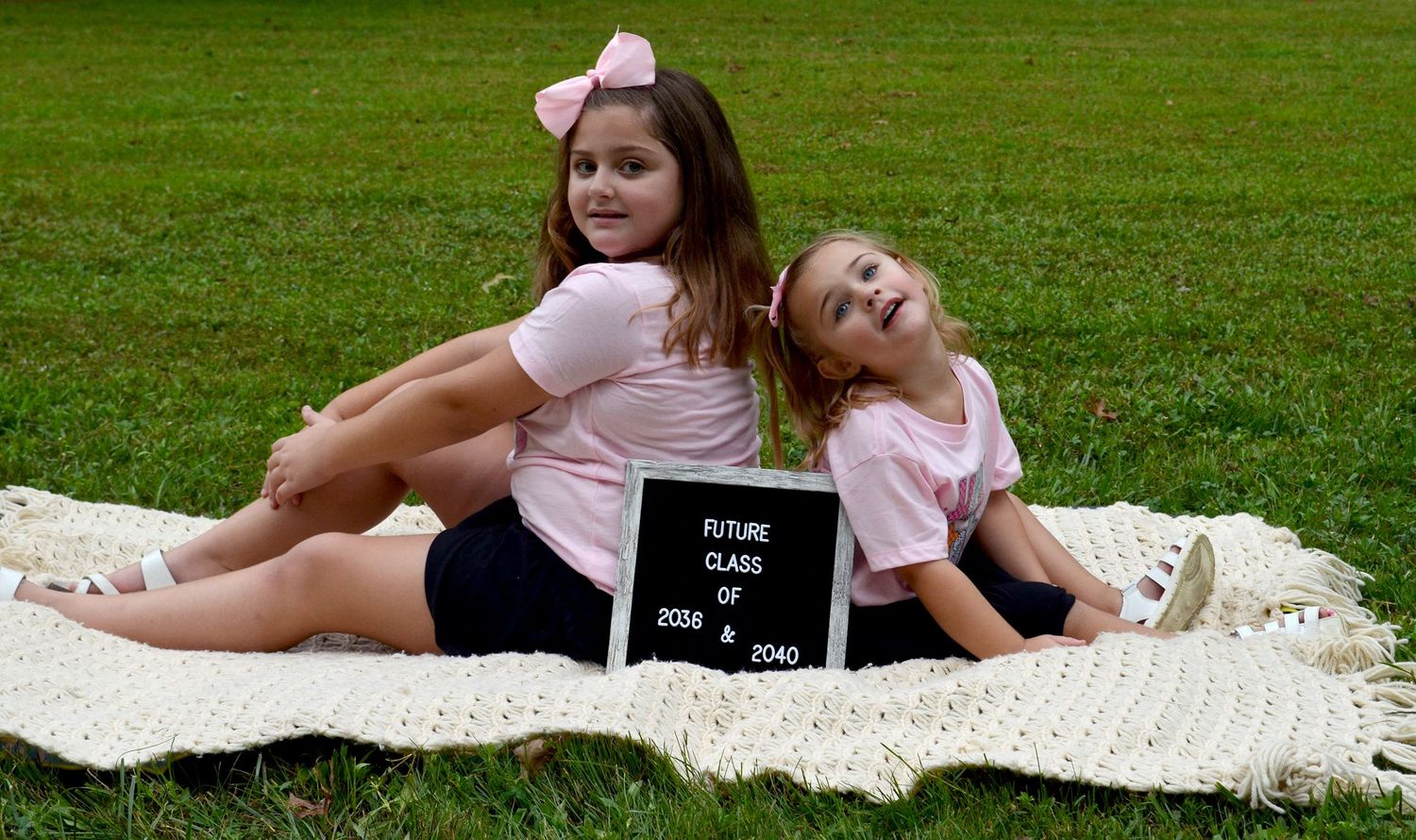 Two young girls in matching pink shirts sit back-to-back on a blanket with a Future Class of 2034 & 2040 sign.