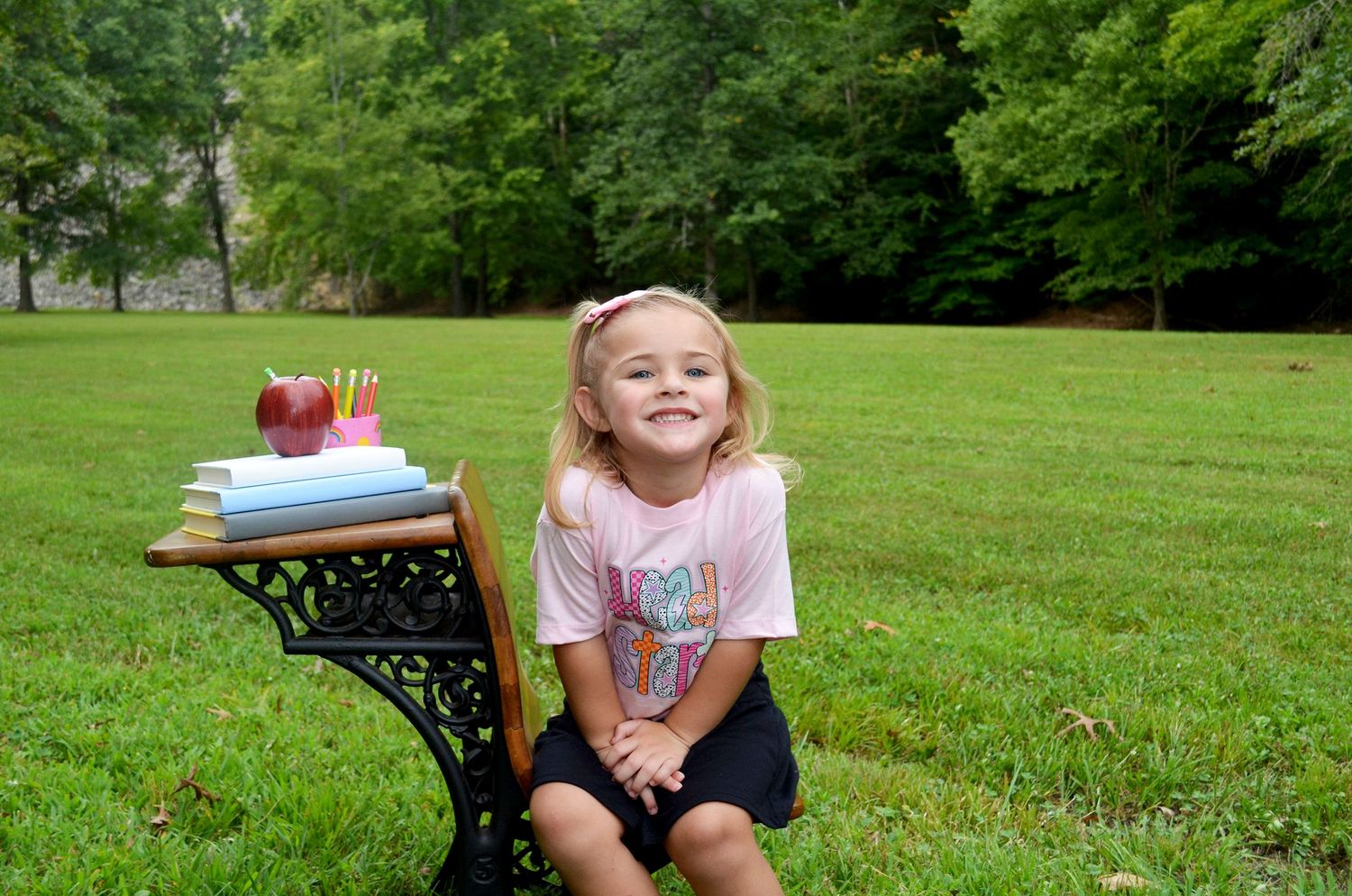 Young girl smiling outdoors beside a school desk with books, pencils, and an apple on green grass.