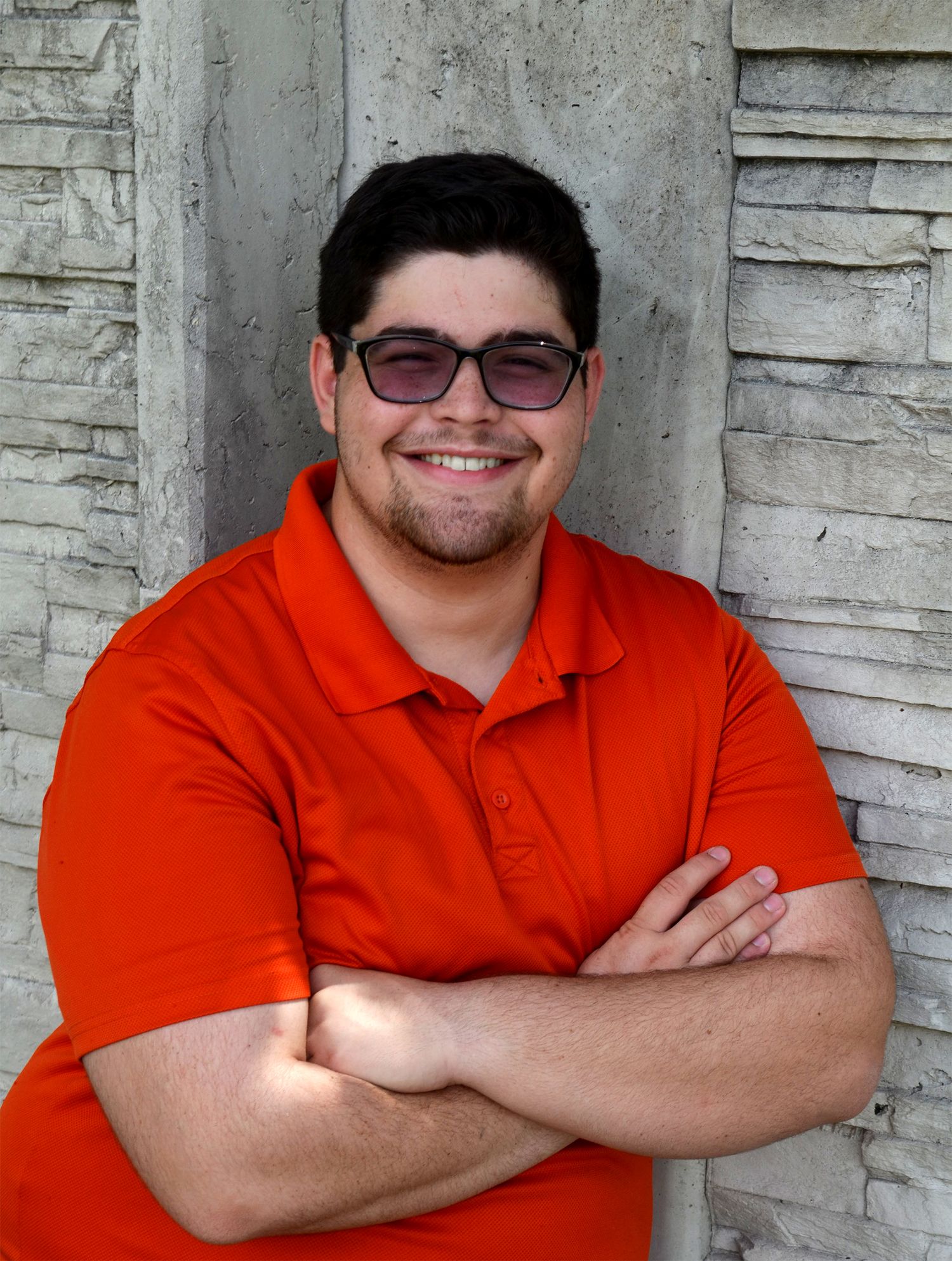 Young man in orange polo shirt with crossed arms and sunglasses smiling against a gray wall.