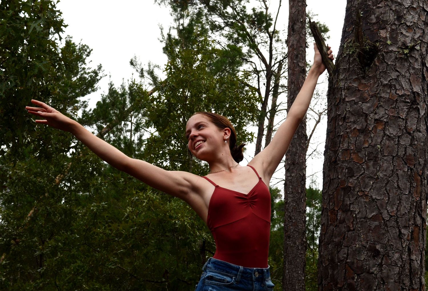 Ballet dancer posing in pine forest with arms outstretched.