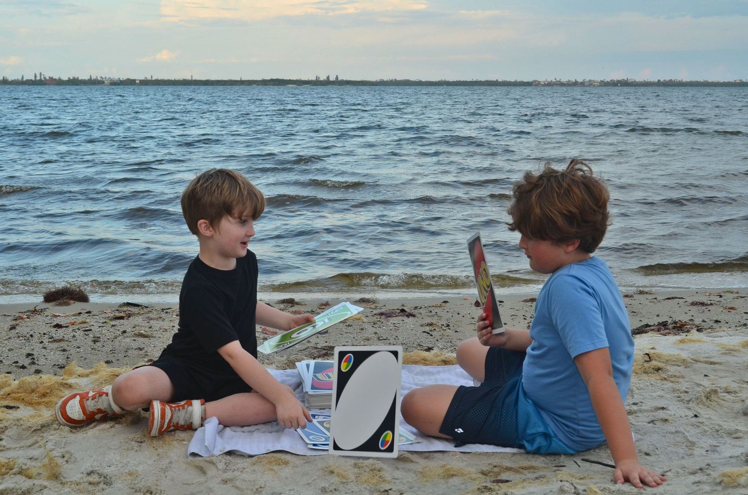 Brothers playing Uno on a beach with water in background.