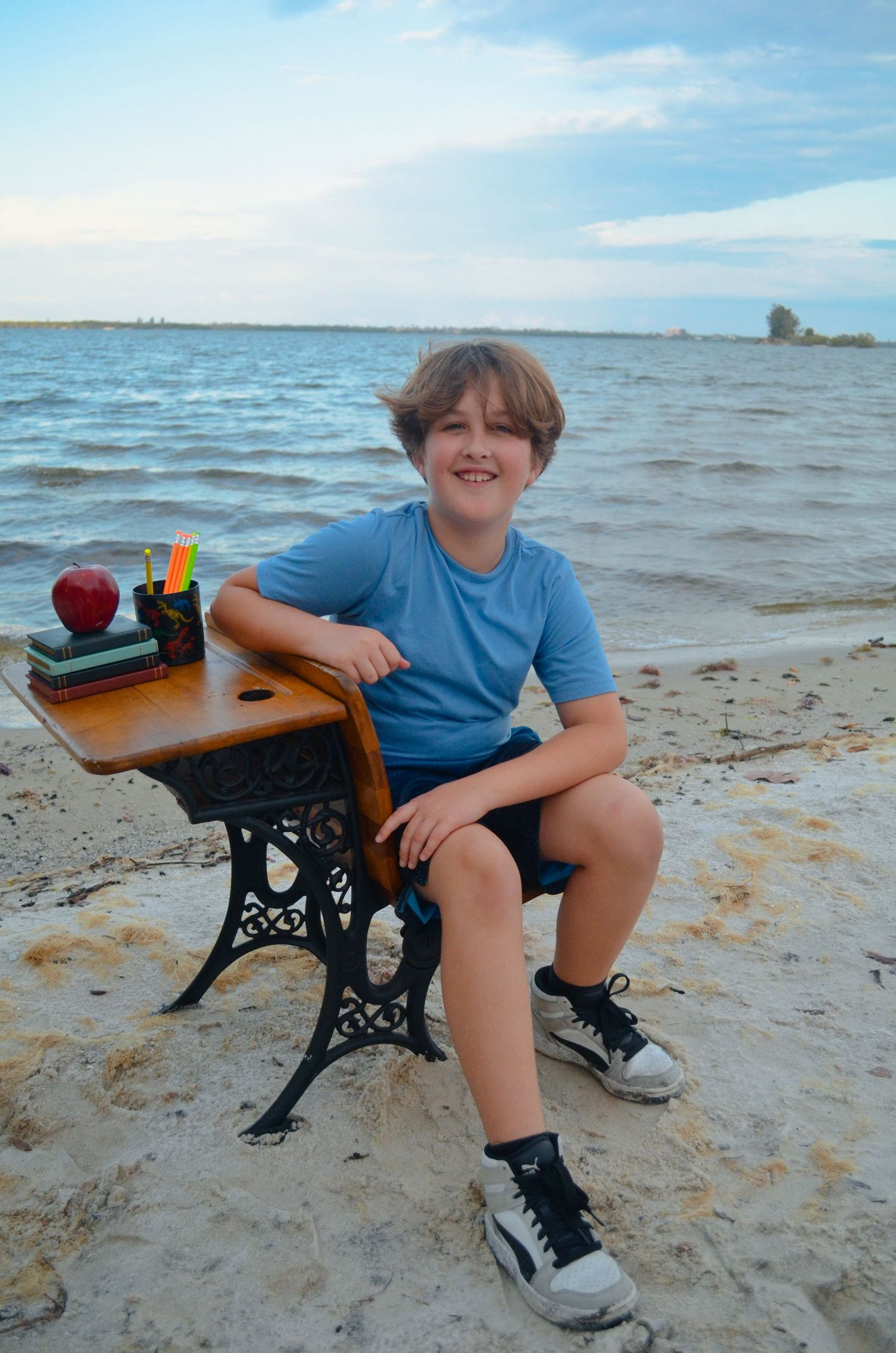 Boy sitting at vintage school desk on sandy beach with ocean backdrop, smiling, wearing blue shirt and sneakers.