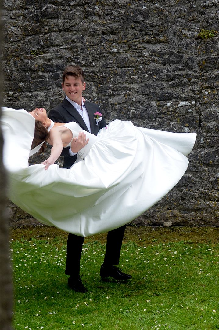 Groom spinning bride on grass beside stone wall.