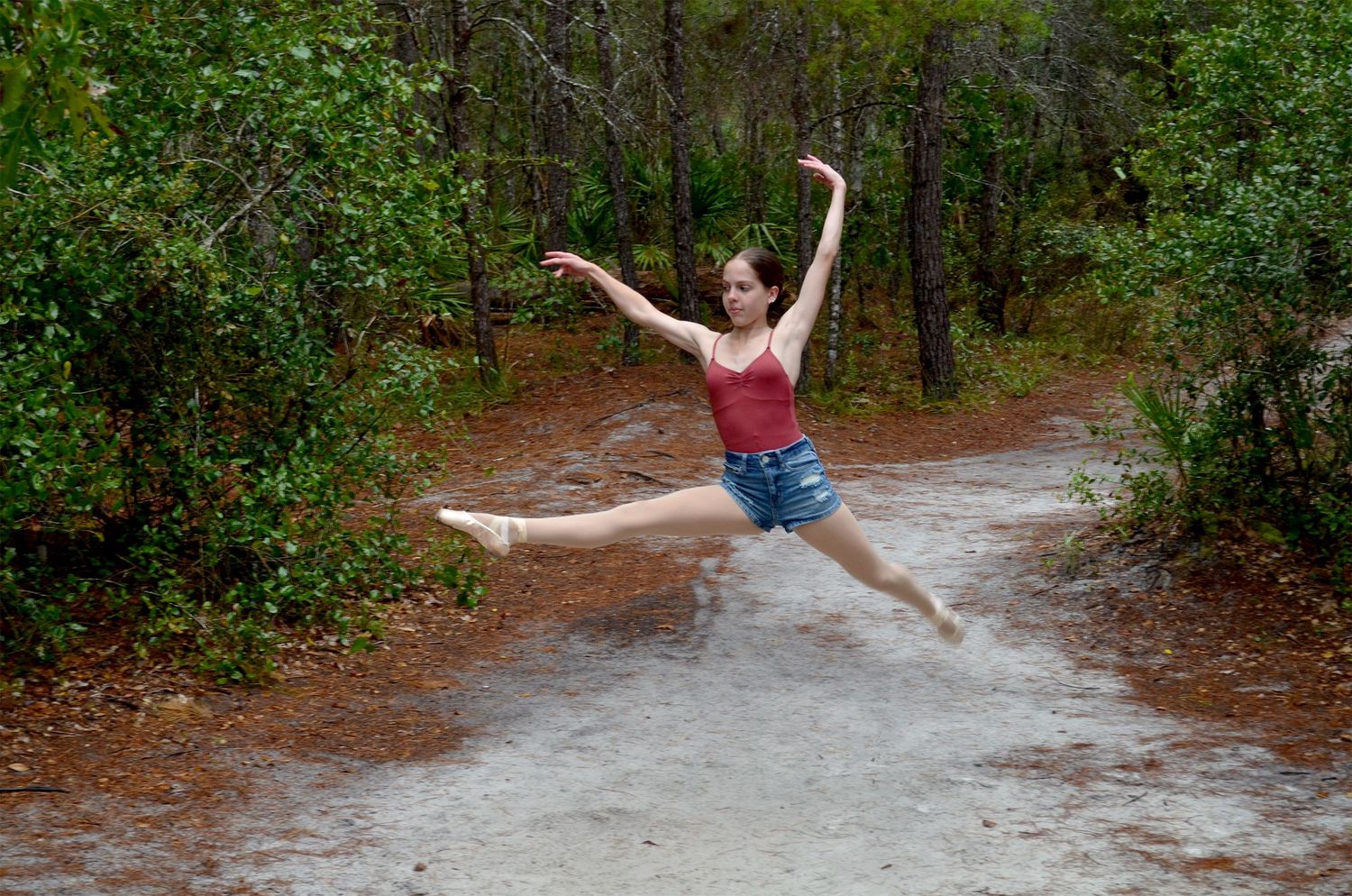 Ballet dancer performing a grand jeté along a sandy forest path.