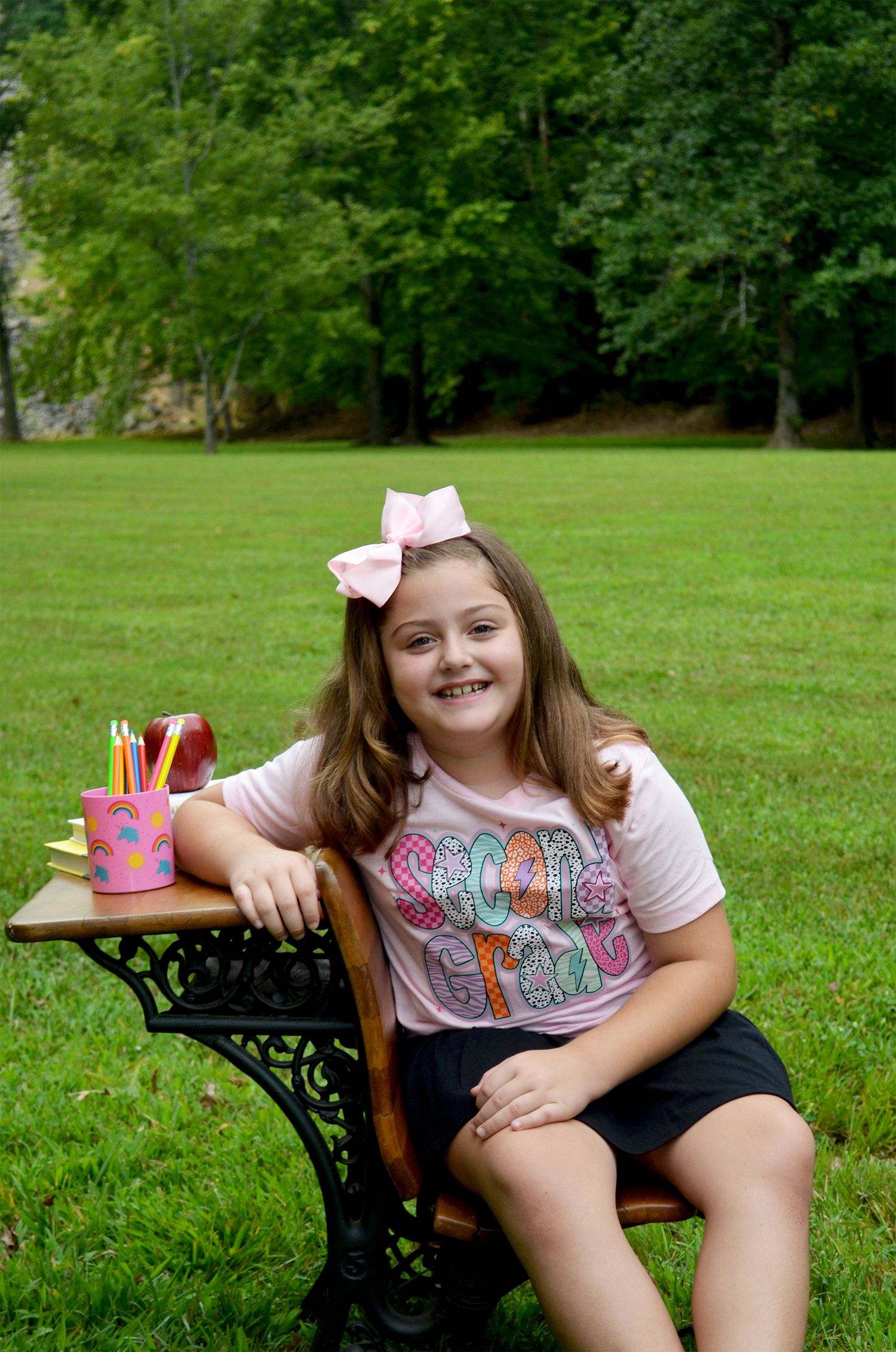 A smiling girl in a pink shirt and bow sits at an antique school desk outdoors on green grass with trees behind her.