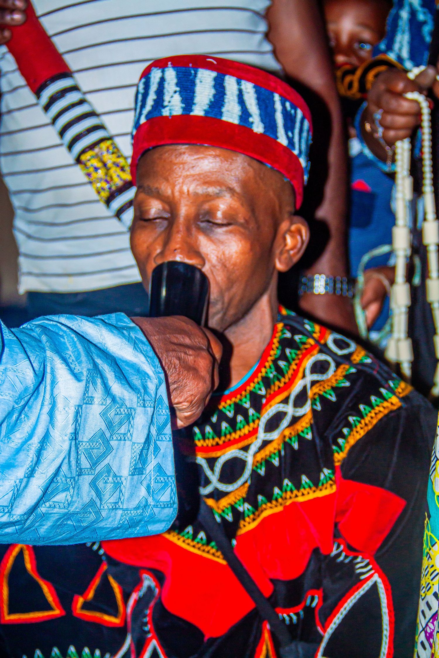 African elder in colorful traditional attire and beaded hat drinking from a ceremonial cup during cultural celebration.