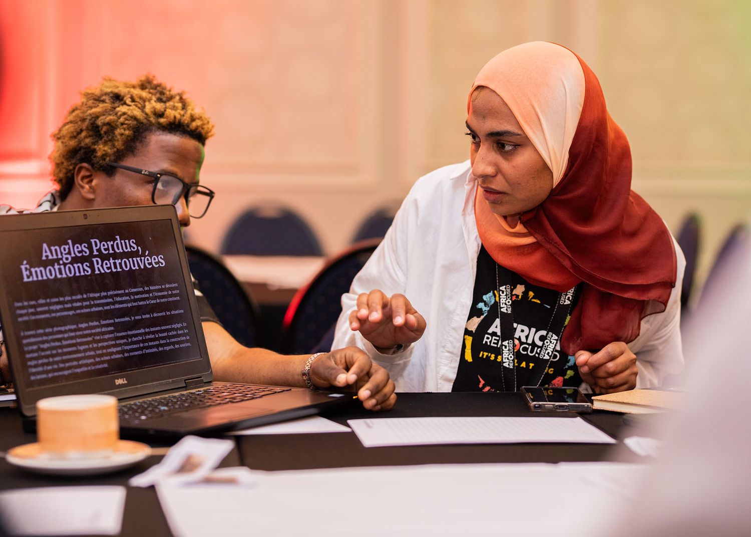 Two women collaborate over a laptop displaying French text at an indoor conference or workshop.